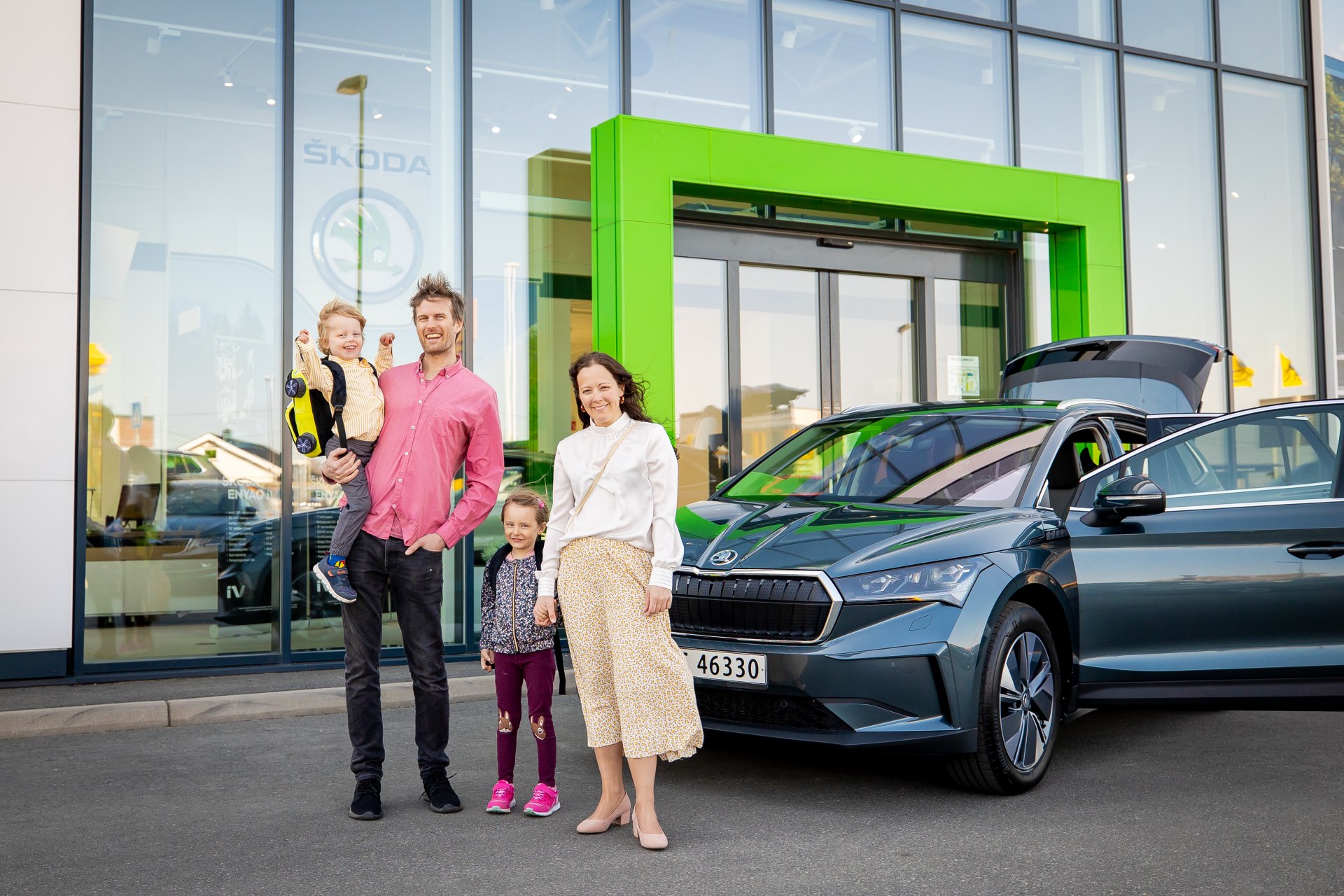 Használt elektromos autó átvétele: család a frissen átvett Škoda Enyaq iV mellett a márkakereskedés előtt